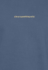 Tissu bleu avec petit texte jaune brodé centré sur la poitrine indiquant "visez quelque chose de solide".