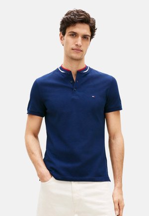 Hombre con una camisa polo de manga corta azul marino con un detalle en el cuello en rojo, blanco y azul, y pantalones de color claro, de pie con una mano en el bolsillo.