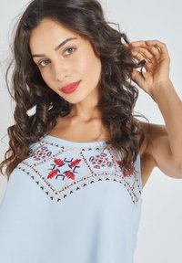 Top sin mangas de color azul claro con patrones florales bordados en rojo y azul marino. Tela suave con un ajuste relajado. Cuello redondo simple.