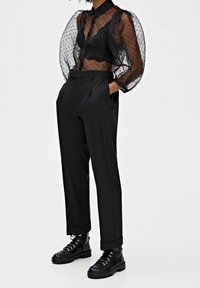 Zwarte doorschijnende blouse met stippenpatroon, gecombineerd met hoge zwarte broeken. Het model draagt zwarte veterschoenen met een dikke zool.