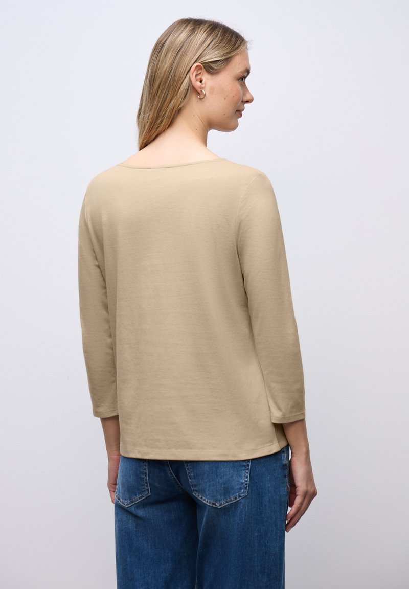 Camicia beige a maniche lunghe con vestibilità comoda, scollatura rotonda e una consistenza liscia e morbida. Abbinata a jeans blu, che mostrano uno stile informale.