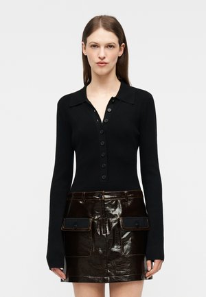 Ženska, oblečena v črno bluzo z dolgimi rokavi in gumbi ter sijočo temno rjavo mini krilo z žepi na sprednji strani, stoji pred enobarvnim ozadjem.