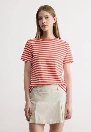 Jonge vrouw die een oranje-wit gestreept T-shirt en een beige geplooide rok draagt, staand tegen een effen witte achtergrond.