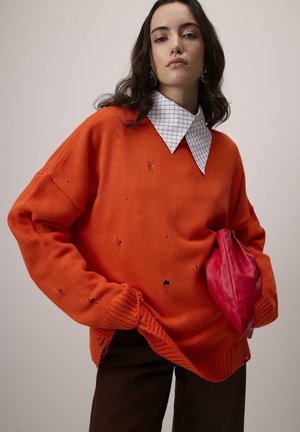 Oranje gebreide trui met versleten gaten, geribbelde manchetten en een wit geruit kraagje. Gepaard met een bruine broek en een roze tas.