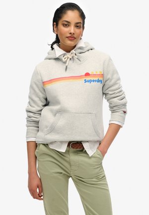 Vrouw draagt een lichtgrijze Superdry hoodie met zonsondergang- en palmboomdesign, een wit shirt eronder en lichtgroene broek met een gevlochten riem.