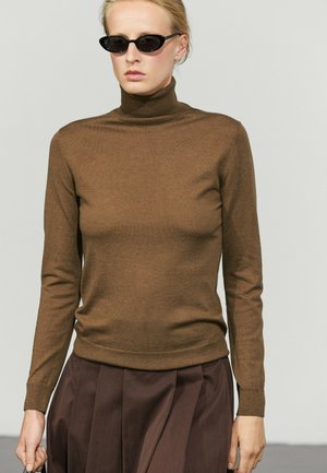 Donna con occhiali da sole neri a forma di occhi di gatto, maglione marrone a collo alto e gonna plissettata marrone scuro su uno sfondo chiaro uniforme.