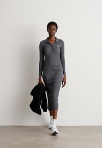 Sivá, dlhá, rukávová, golierová šaty s úzkym strihom a dĺžkou po kolená. Modelka drží čiernu bundu s hladkým povrchom. Biele športové topánky.