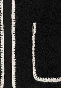 Schwarzer Fleece-Stoff mit weißen Nähten an den Kanten, ausgestattet mit einer aufgesetzten Tasche. Die Textur ist weich und strukturiert, mit einem gemütlichen Aussehen.