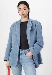 Svetlo modra denim jakna velikosti oversized z izrezanimi klapami, z enim gumbom in žepom na prsih, v kombinaciji z črnim puloverjem in svetlo modrimi jeans hlačami.