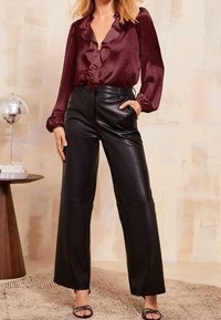 Blouse en soie bordeaux avec un col à volants et pantalon large en cuir noir, doté de poches latérales, porté avec des sandales à brides sur un tapis texturé.