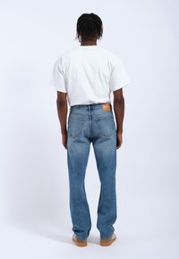 Jeans in denim azzurro chiaro con taglio dritto, con tasche posteriori e una toppa in pelle marrone in vita, abbinati a una maglietta bianca.