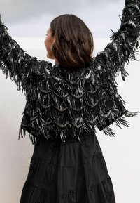 Zwarte cropped jas met kralen en franjes, getextureerd met gelaagde paillettenpatronen, gedragen over een zwarte jurk met lagen.