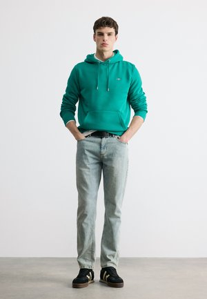 Νέος άνδρας στέκεται με τα χέρια στις τσέπες, φοράει μια τυρκουάζ μπλούζα με κουκούλα, ανοιχτόχρωμα μπλε τζιν και μαύρα αθλητικά παπούτσια, μπροστά σε ένα απλό λευκό φόντο.