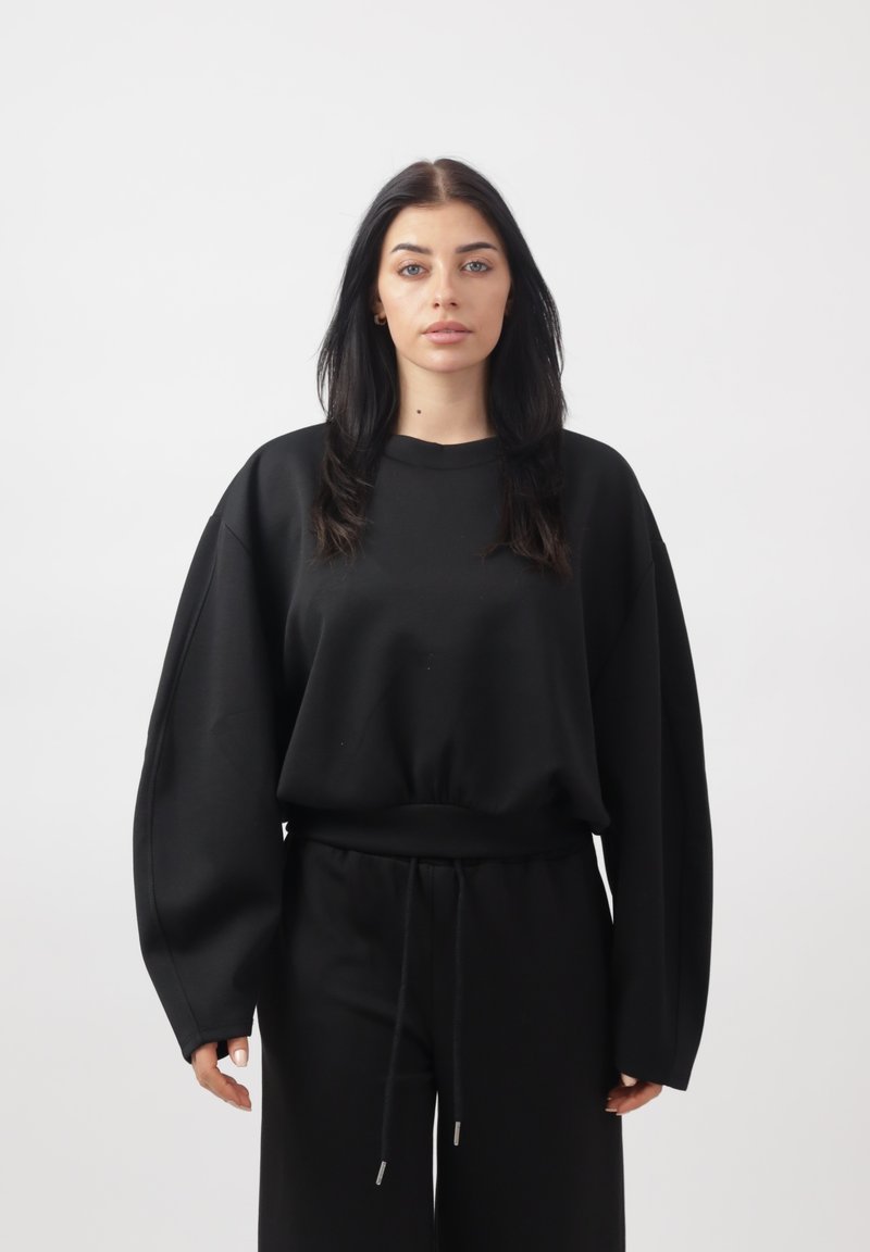 Donna con capelli lunghi e scuri che indossa una felpa nera larga e pantaloni della tuta neri abbinati, in piedi davanti a uno sfondo chiaro uniforme.