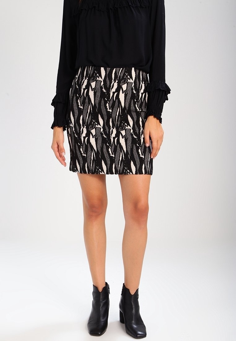 Femme portant un haut noir à manches longues, une minijupe noire et blanche à motifs, et des bottines noires, debout devant un fond uni.