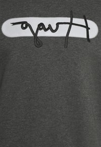 Camiseta de algodón gris con un parche horizontal blanco que lleva texto en cursiva negra. La tela es suave con un acabado ligeramente texturizado.