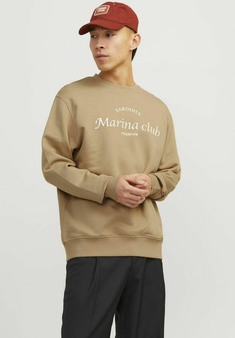 Khaki sweatshirt with white text "Sardinia Marina club PREMIUM" on front. Ribbed cuffs and hem. Paired with dark trousers and a red cap.