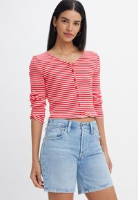 Röd- och vitrandig, långärmad kort cardigan med rund halsringning och knappstängning, tillsammans med ljusblå denimshorts.