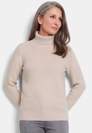 Frau mit grauen und braunen Haaren, die einen hellbeigen gestrickten Rollkragenpullover und graue Hosen trägt, steht mit den Händen hinter dem Rücken.