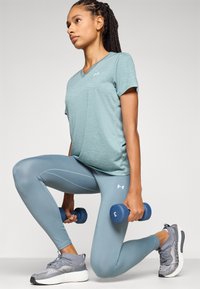 Világoskék sport póló, kék leggings, szürke sportcipő, kék kézi súlyzók a kezében. Kiemelkedő logó a pólón és a leggingsen, sima tapintású.