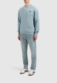 Light blue sweatshirt with crew neckline and ribbed cuffs, featuring a small black logo on the chest. Paired with light blue jeans and sneakers.