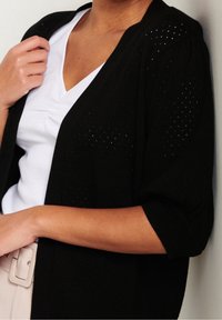 Kobieta w czarnej, fakturowanej kardiganie na białej bluzce z wycięciem w serek, z widocznym beżowym paskiem, pozująca z ręką przy ramieniu i ramieniem opartym na talii.