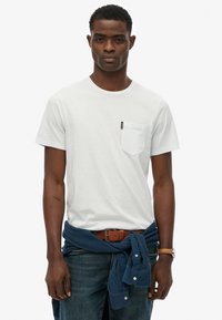 Witte T-shirt met korte mouwen en een borstzak, gecombineerd met blauwe jeans en een marineblauw overhemd dat om de taille is vastgebonden. Bruine riem en horloge.