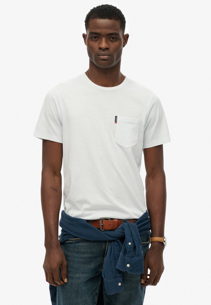 Witte T-shirt met korte mouwen en een borstzak, gecombineerd met blauwe jeans en een marineblauw overhemd dat om de taille is vastgebonden. Bruine riem en horloge.