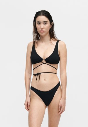 Γυναίκα με βρεγμένα μαλλιά, που φοράει μαύρο μπικίνι με φαρδιά κορδόνια και ταιριαστό κάτω μέρος, στέκεται μπροστά σε λευκό απλό φόντο.