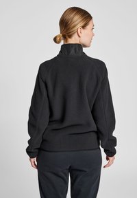 Svart fleece-pullov med hög krage, strukturerad tyg, raglanärmar och kontrasterande slät krage. Bärs med mörka matchande byxor.