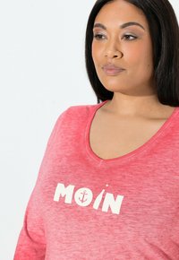 Lyserød V-hals shirt med en let struktureret stof, der har hvid tekst "MOIN" og et ankersymbol. Designet er afslappet og uformelt.