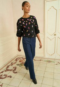 Melnā džemperī ar krāsainiem ziedu izšuvumiem, apvienots ar tumši ziliem platām biksēm. Modelis valkā melnas augstpapēžu kurpes, pozējot flīzētā telpā.