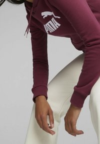 Burgunder sweatshirt med hvid "PUMA" logo og kattografik, med snøre i huen og ribbede manchetter, parret med off-white bukser.