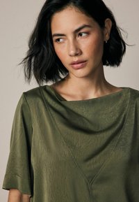 Olijfgroene blouse met een gedrapeerde halslijn, korte mouwen en een textuurstof. Kenmerkt zich door een subtiele glans en geometrische naaddetails.