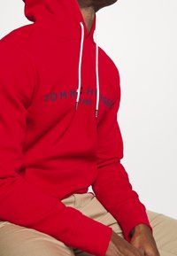 Homme assis portant un sweat à capuche rouge Tommy Hilfiger avec des cordons blancs et un pantalon beige, sur un fond blanc uni.