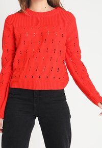 Červený pletený svetr s texturovaným vzorem a průhlednými detaily, žebrovaným výstřihem a dlouhými rukávy. Kombinován s černými kalhotami.