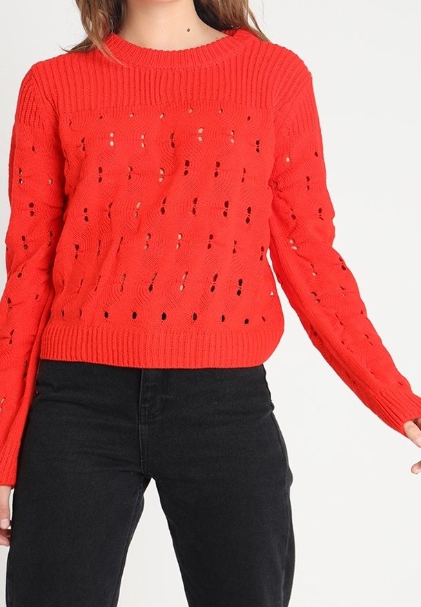 Červený pletený svetr s texturovaným vzorem a průhlednými detaily, žebrovaným výstřihem a dlouhými rukávy. Kombinován s černými kalhotami.