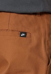 Los pantalones de tela marrón de Nike cuentan con un bolsillo trasero que tiene una pequeña etiqueta de logo negra. La textura es suave con un acabado en color sólido.