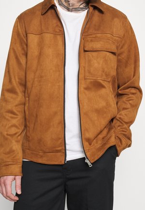 Mężczyzna w jasnobeżowej zamszowej kurtce na zamek błyskawiczny, pod nią biała koszulka, czarne spodnie i widoczne tatuaże na szyi.