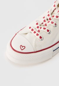 Λευκό καμβά sneaker με κορδόνια με κόκκινα σχέδια καρδιάς, κόκκινο καουτσούκ πέλμα στα δάχτυλα και ρίγες στη σόλα. Διαθέτει μια μικρή λεπτομέρεια καρδιάς στα δάχτυλα.