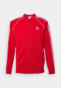 Röd zip-up träningsjacka i mjukt material, med vita randiga detaljer längs axlarna och armarna samt en broderad logotyp på bröstet.