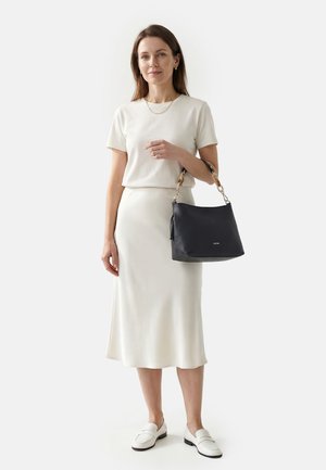 Mujer con top de manga corta blanco y falda, sosteniendo un bolso negro con asa de carey, de pie sobre un fondo blanco liso.