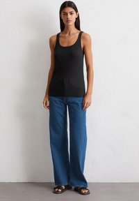 Czarny ryflowany top na ramiączkach z szerokimi szelkami, zestawiony z luźnymi niebieskimi dżinsami. Modelka nosi płaskie czarne sandały.