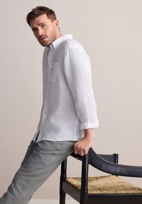 Camicia di lino bianca con maniche arrotolate e una tasca sul petto, abbinata a pantaloni grigi, appoggiata contro una sedia in legno nero con seduta intrecciata.