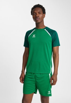 Man wearing green hummel sports jersey and matching shorts, standing against a plain light background.