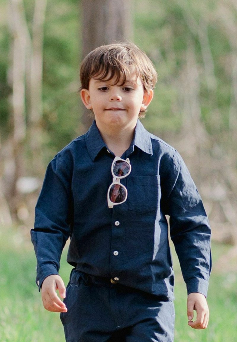 Napapaita ja housut syvän sinisestä kankaasta, jossa on etutasku. Lapsi, jolla on tyylikkäät aurinkolasit, kävelee nurmikolla luonnollisessa ympäristössä.