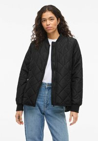 Črna prešita bomber jakna z zadrgo spredaj in rebrastimi manšetami, kombinirana z belo srajco in svetlim modrim jeansom.