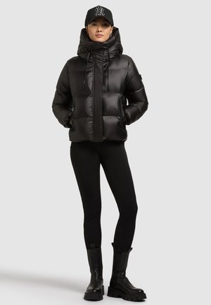 Svart vadderad jacka med luva, glansigt utförande, quiltad design och justerbara dragsnören. Kombinerad med svarta leggings och klumpiga svarta stövlar.