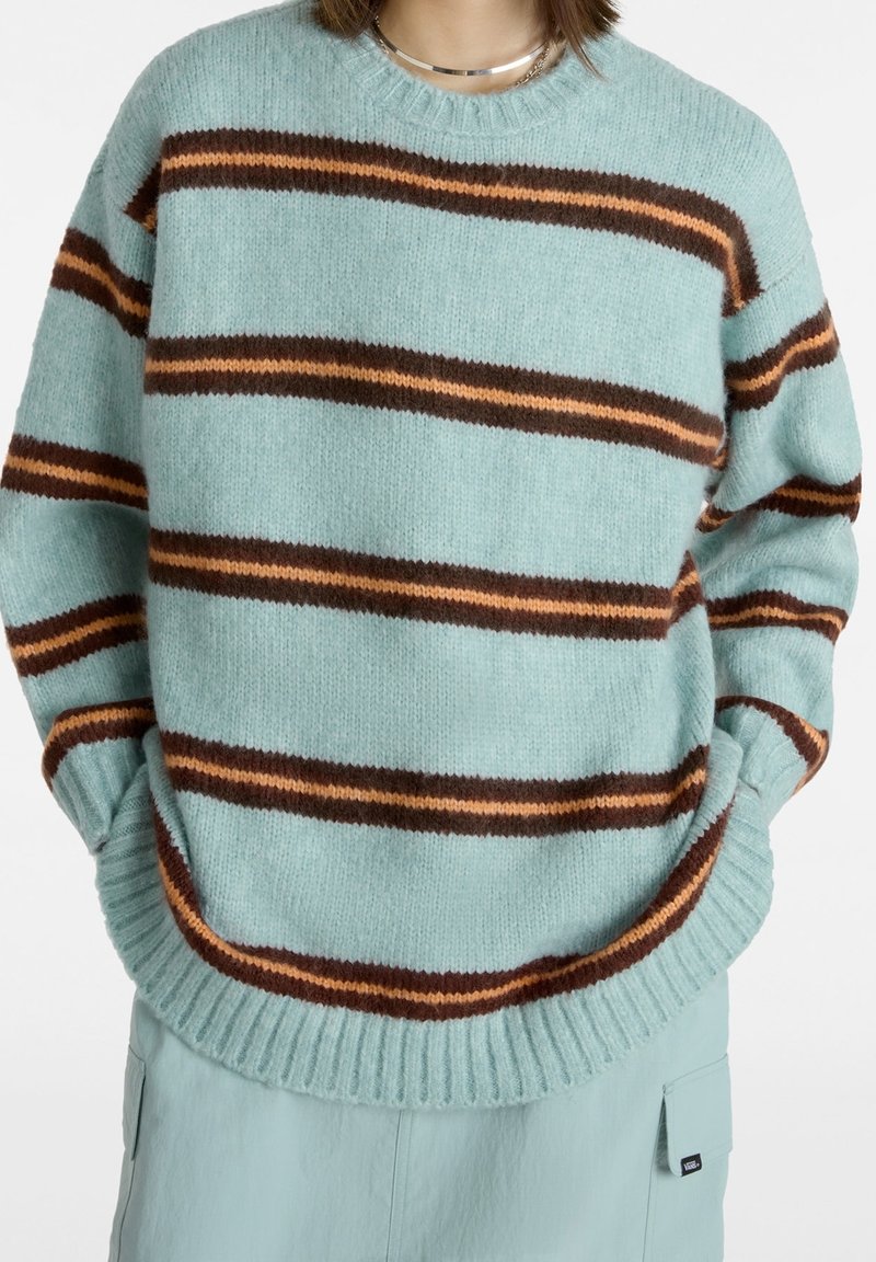 Personne portant un pull en maille bleu clair avec des rayures horizontales marron foncé et orange, assorti à un pantalon bleu clair avec des poches latérales.