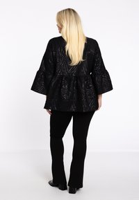 Schwarzes, strukturiertes Oberteil mit Glockenärmeln und Peplum-Taillendetail, aus leichtem Stoff. Kombiniert mit figurbetonten schwarzen Hosen und Stiefeletten mit Absatz.